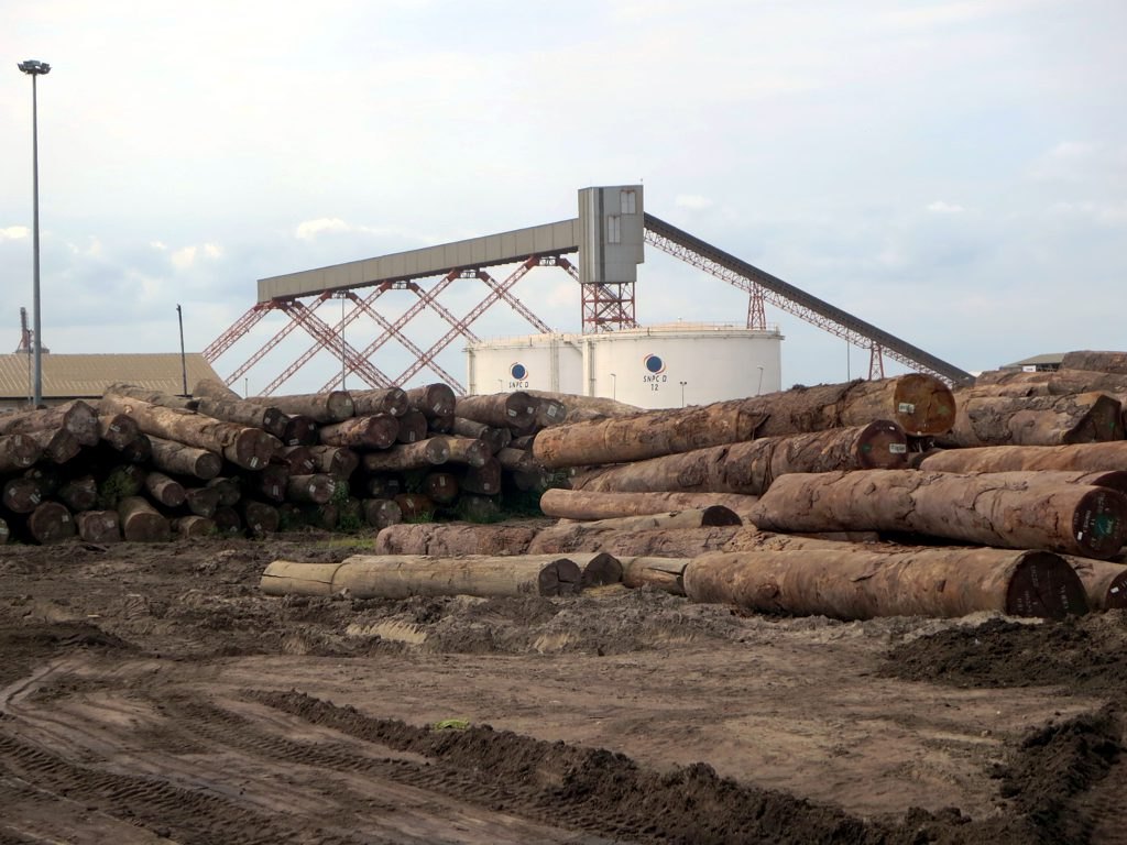 Stacks of tropical logs in front of white oil storage tanks marked SNPC at the port of Pointe-Noire