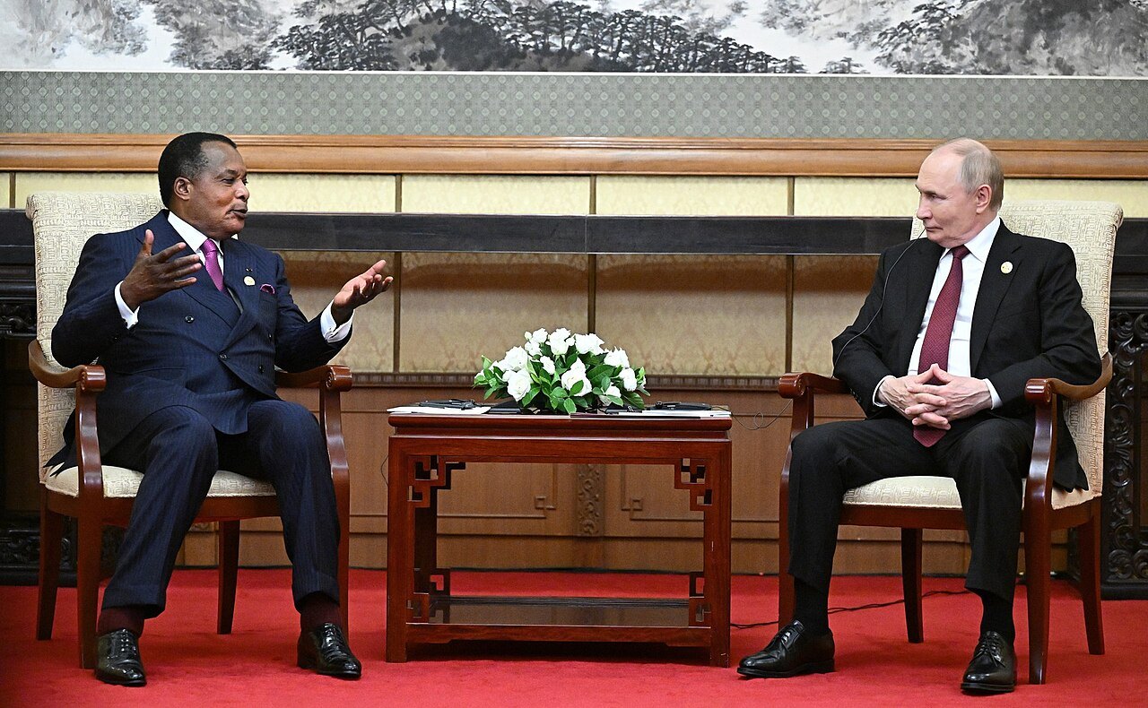 Denis Sassou Nguesso gesturing while seated opposite Vladimir Putin at a meeting room in Beijing