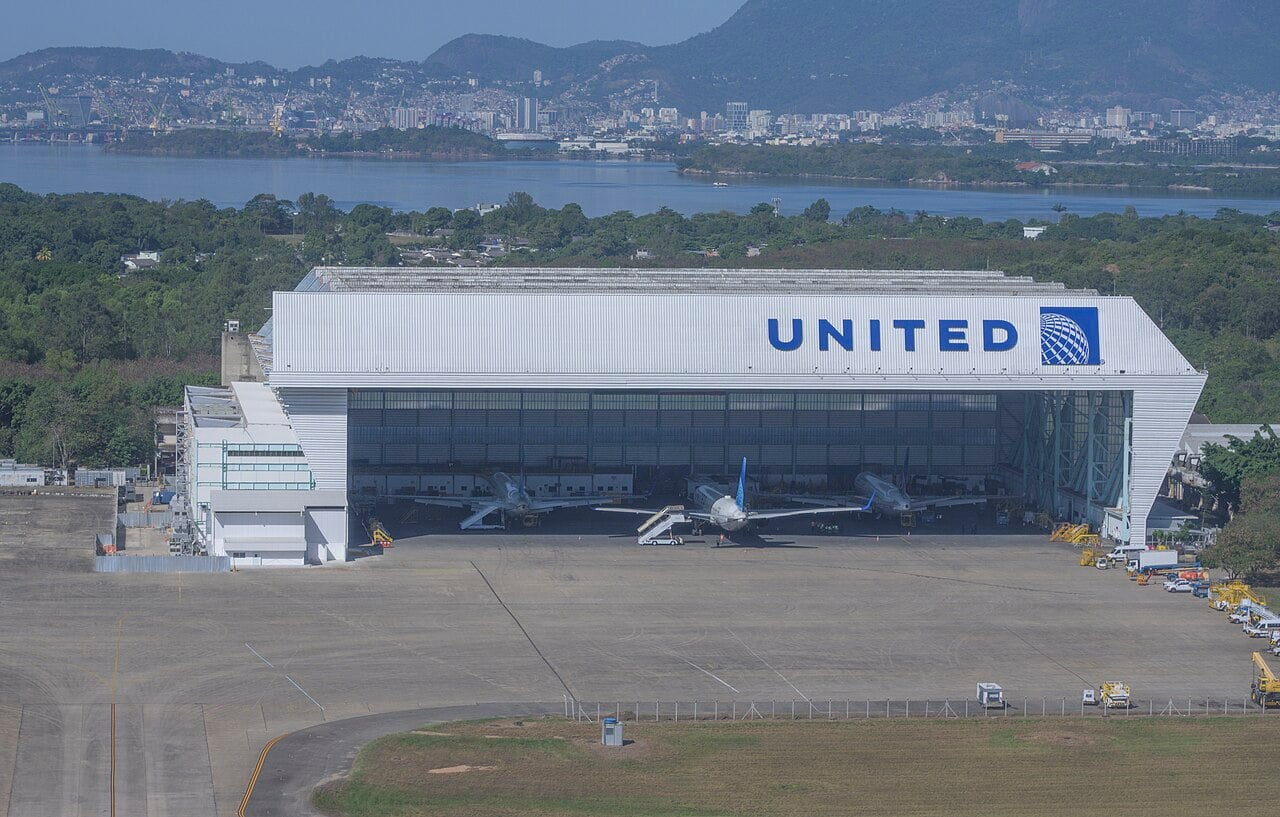 A large white hangar with the United logo, three jets parked inside, at Galeão airport with the water and the city behind