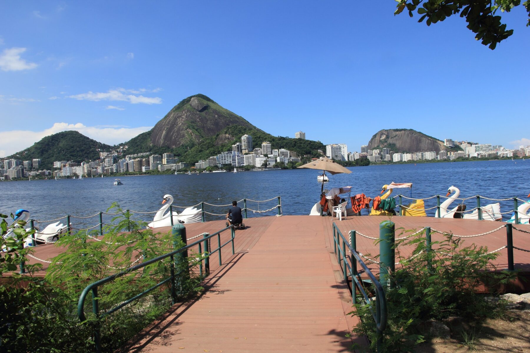 Pedal boats shaped like swans at a jetty on Lagoa Rodrigo de Freitas in Rio de Janeiro, with the Two Brothers hills and Zona Sul apartment blocks across the water.