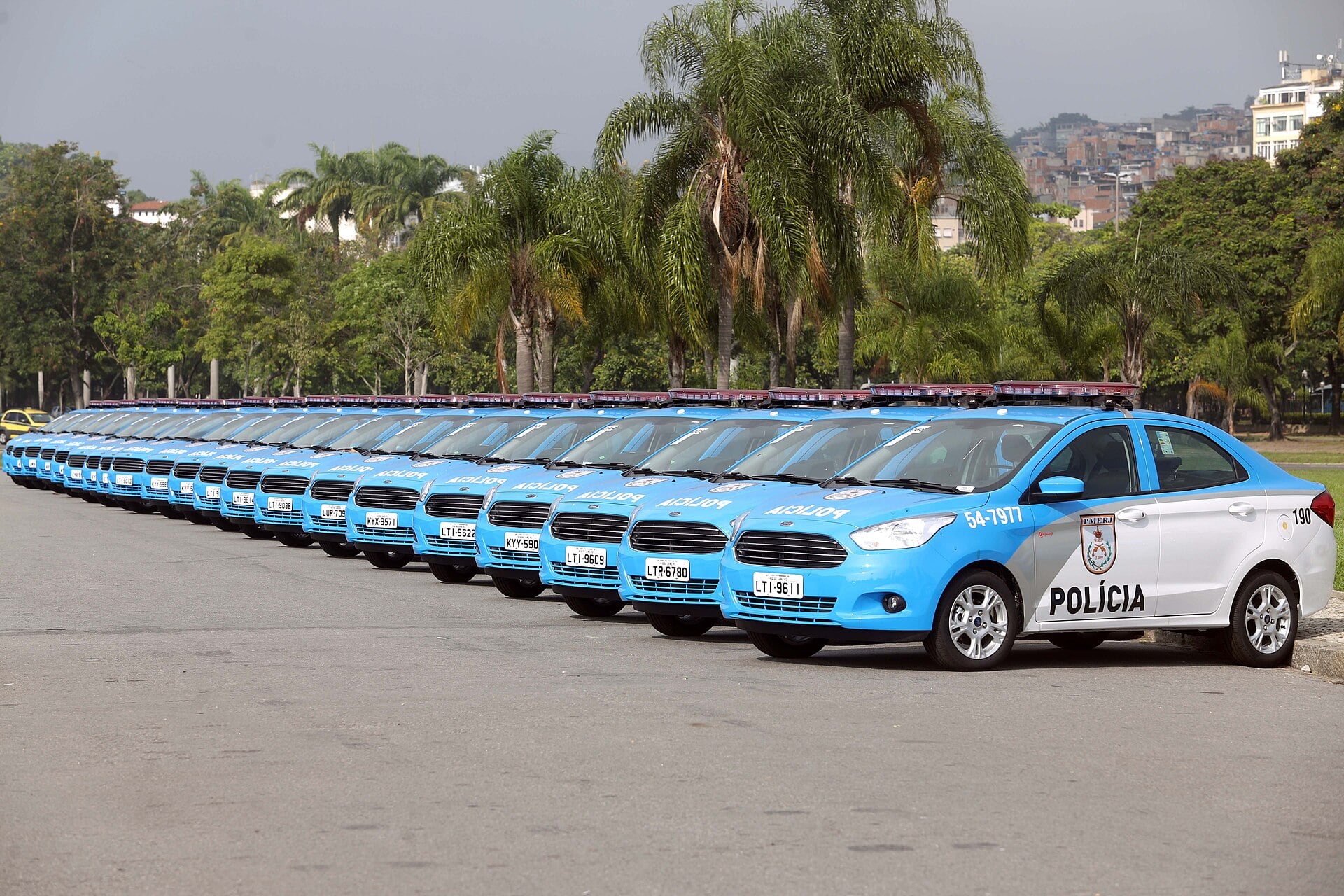A line of blue and white Rio de Janeiro Military Police patrol cars parked along a road beside palm trees