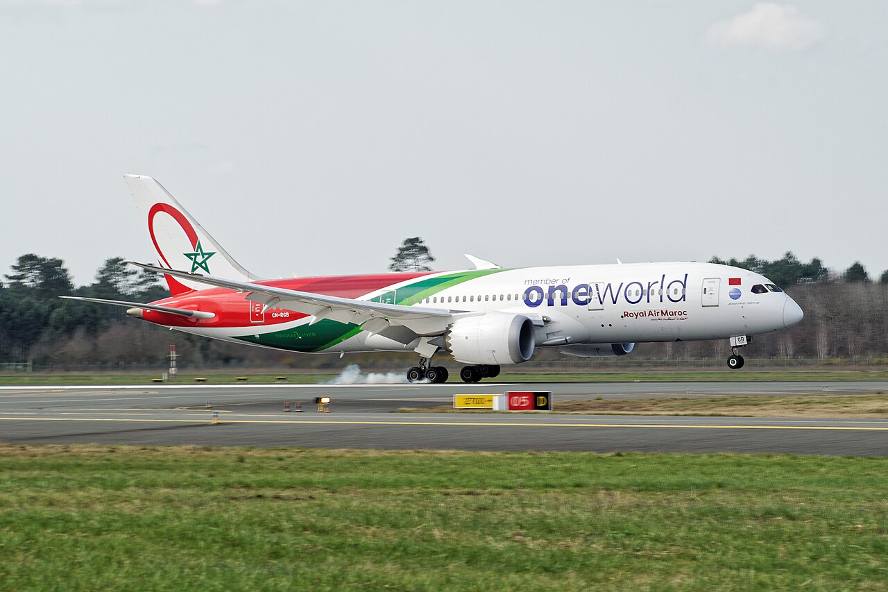 A red, green and white Royal Air Maroc Boeing 787-8 Dreamliner in Oneworld livery touching down on a runway, with a puff of tire smoke.