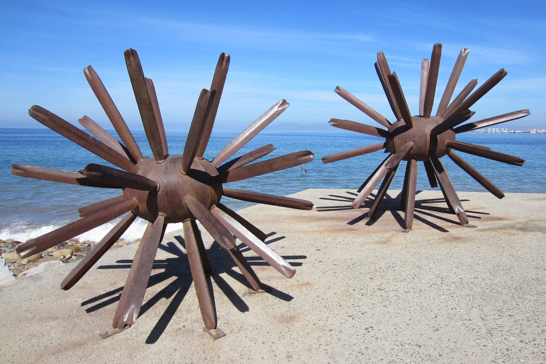 Two large rusty metal sculptures shaped like sea urchins on the paved Malecón seafront in Puerto Vallarta, with the blue sea of Banderas Bay and a clear sky behind