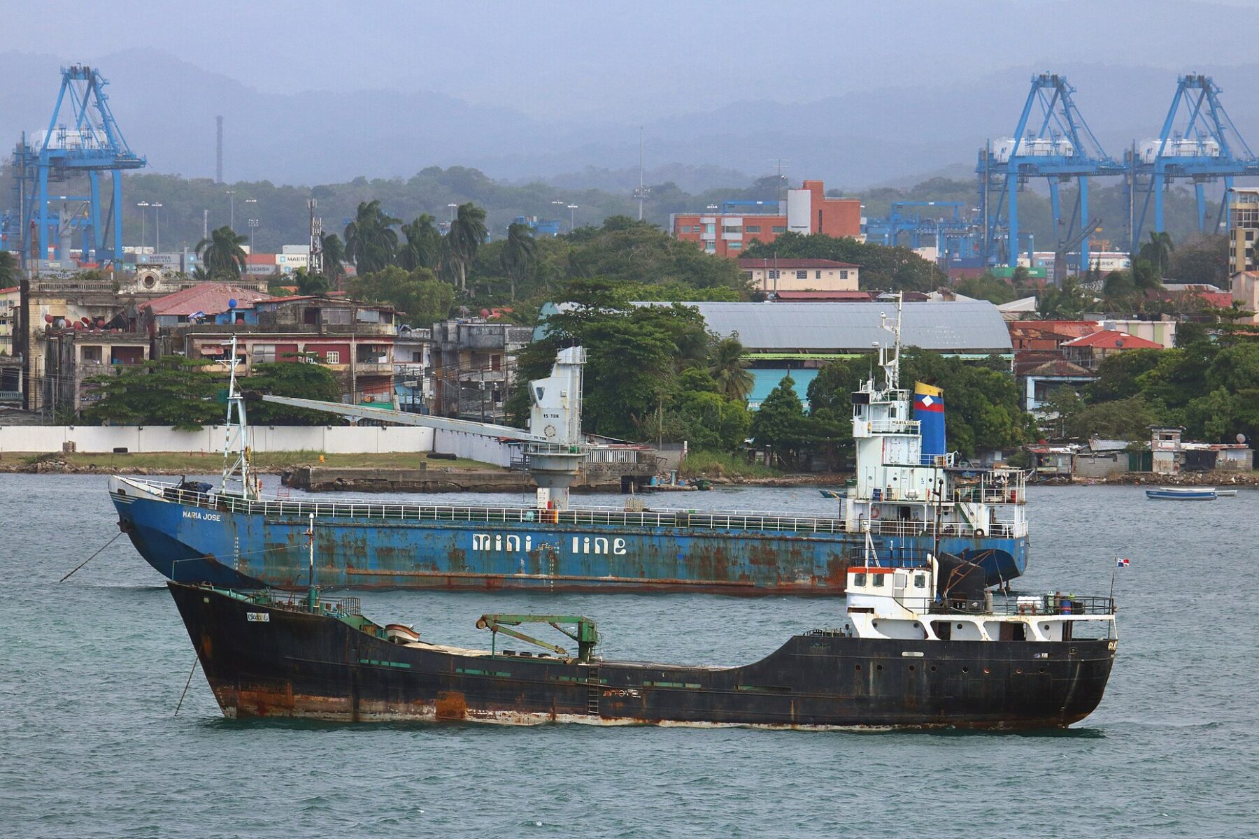 Two rust-streaked general cargo ships at anchor off Colón, Panama, with blue port cranes and green hills behind