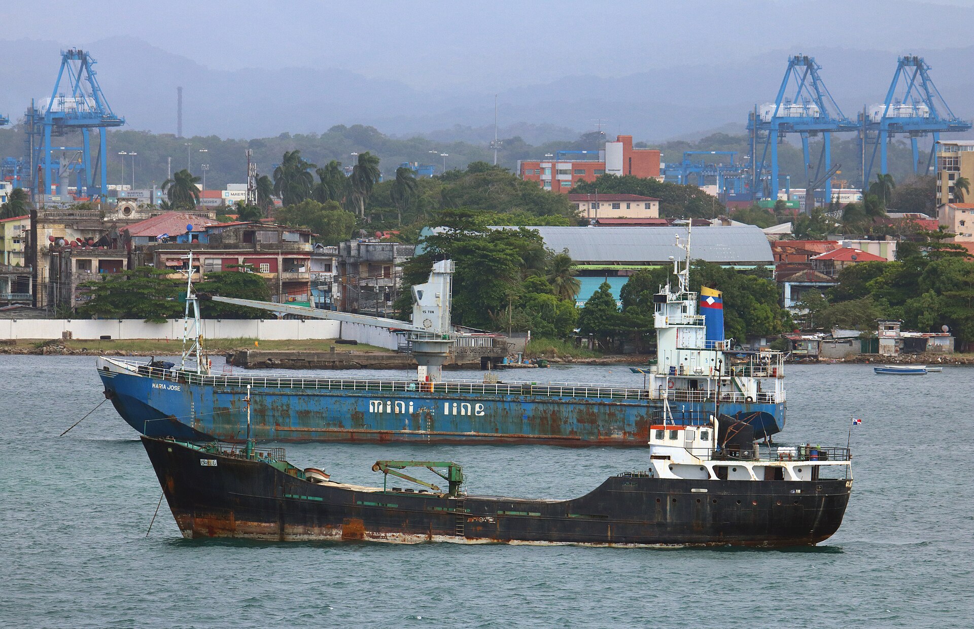 Two rust-streaked general cargo ships at anchor off Colón, Panama, with blue port cranes and green hills behind