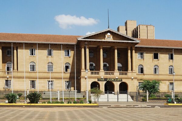 The colonnaded sandstone Judiciary building in Nairobi with a red-tiled roof, flagpoles and a paved forecourt under a blue sky