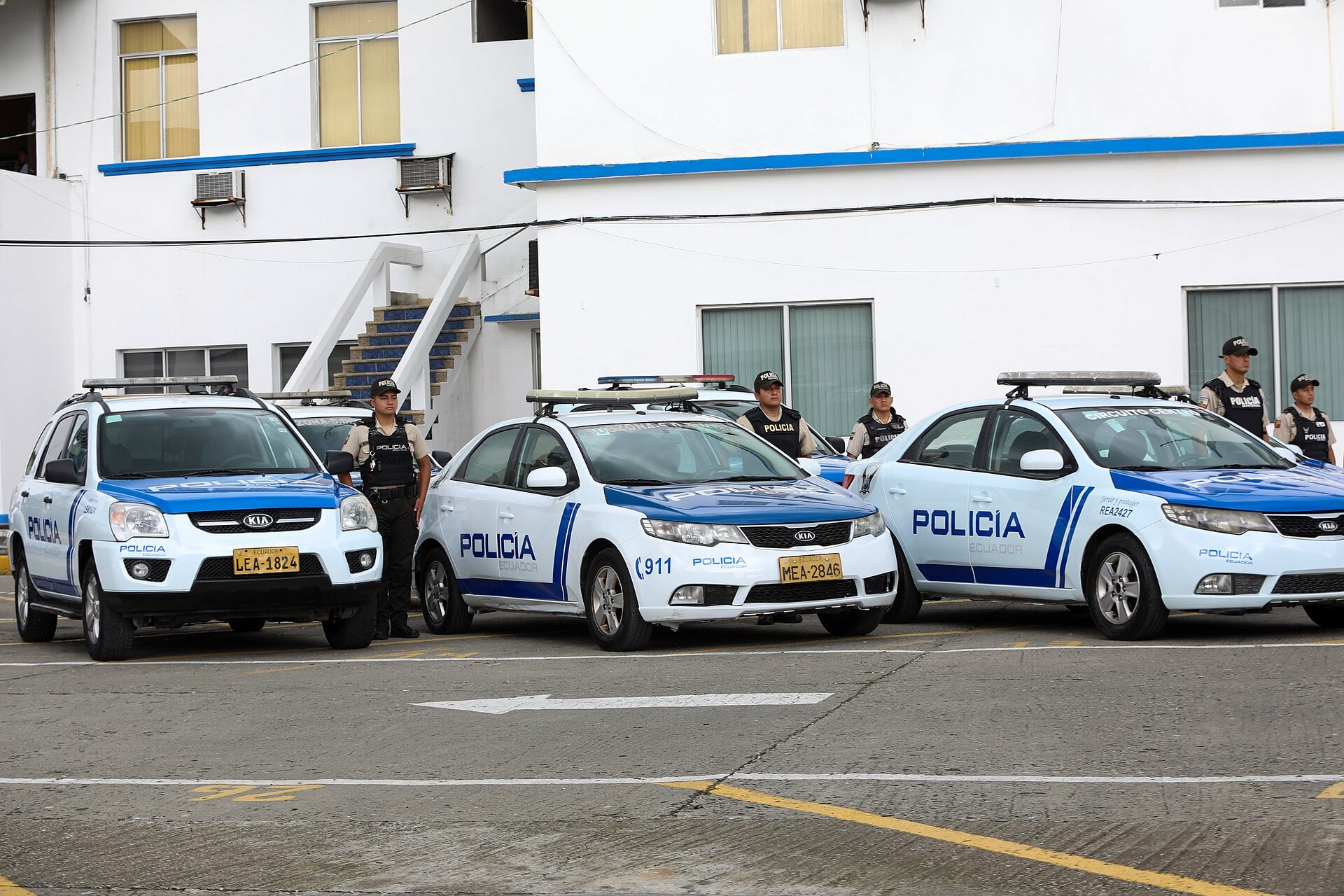 Ecuadorian police officers in vests stand beside white and blue patrol cars marked Policía outside a white building