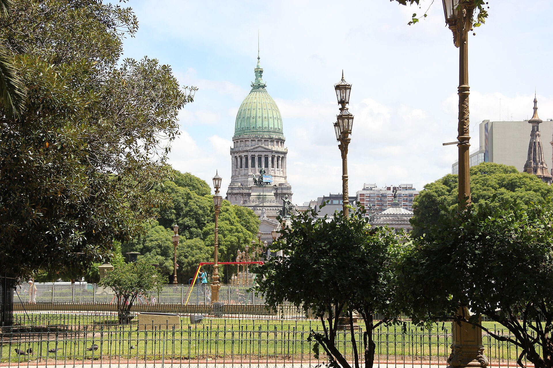 The green dome of Argentina's Palace of the National Congress seen across Plaza del Congreso through trees and ornate street lamps