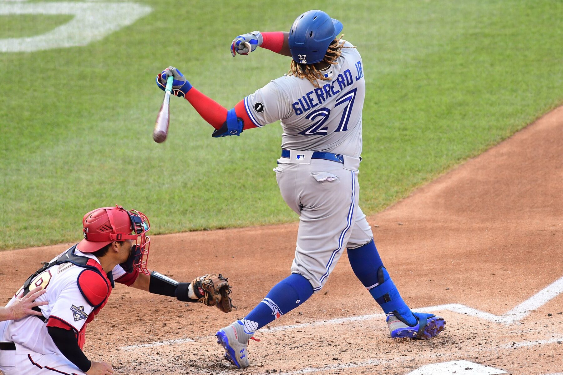 Vladimir Guerrero Jr. in a grey Toronto Blue Jays road uniform with number 27 and a blue helmet follows through on a swing as a catcher in a red helmet crouches behind the plate