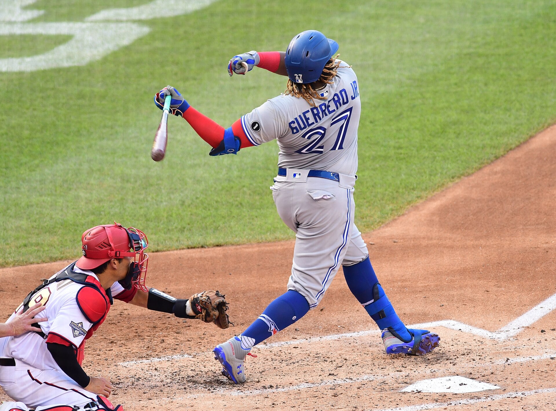 Vladimir Guerrero Jr. in a grey Toronto Blue Jays road uniform with number 27 and a blue helmet follows through on a swing as a catcher in a red helmet crouches behind the plate