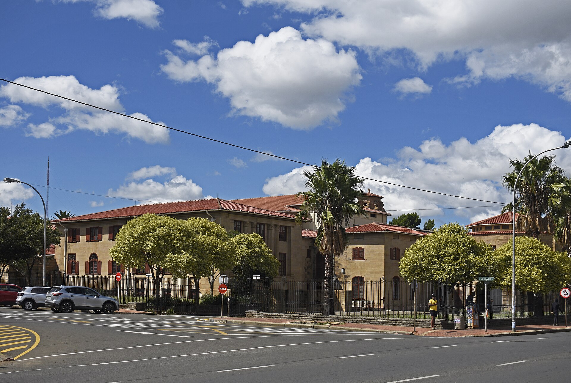 The sandstone Bloemfontein City Hall with red-tiled roofs behind trees and palms at a street corner under a blue sky with white clouds