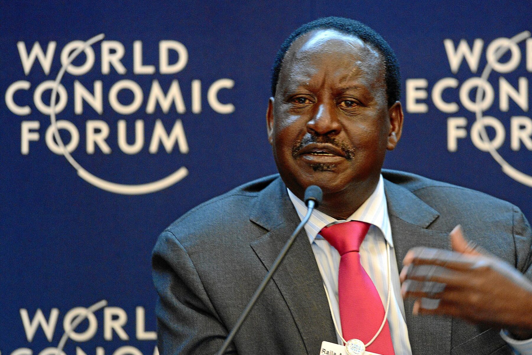 Raila Odinga in a grey suit and red tie speaking into a microphone in front of a blue World Economic Forum backdrop
