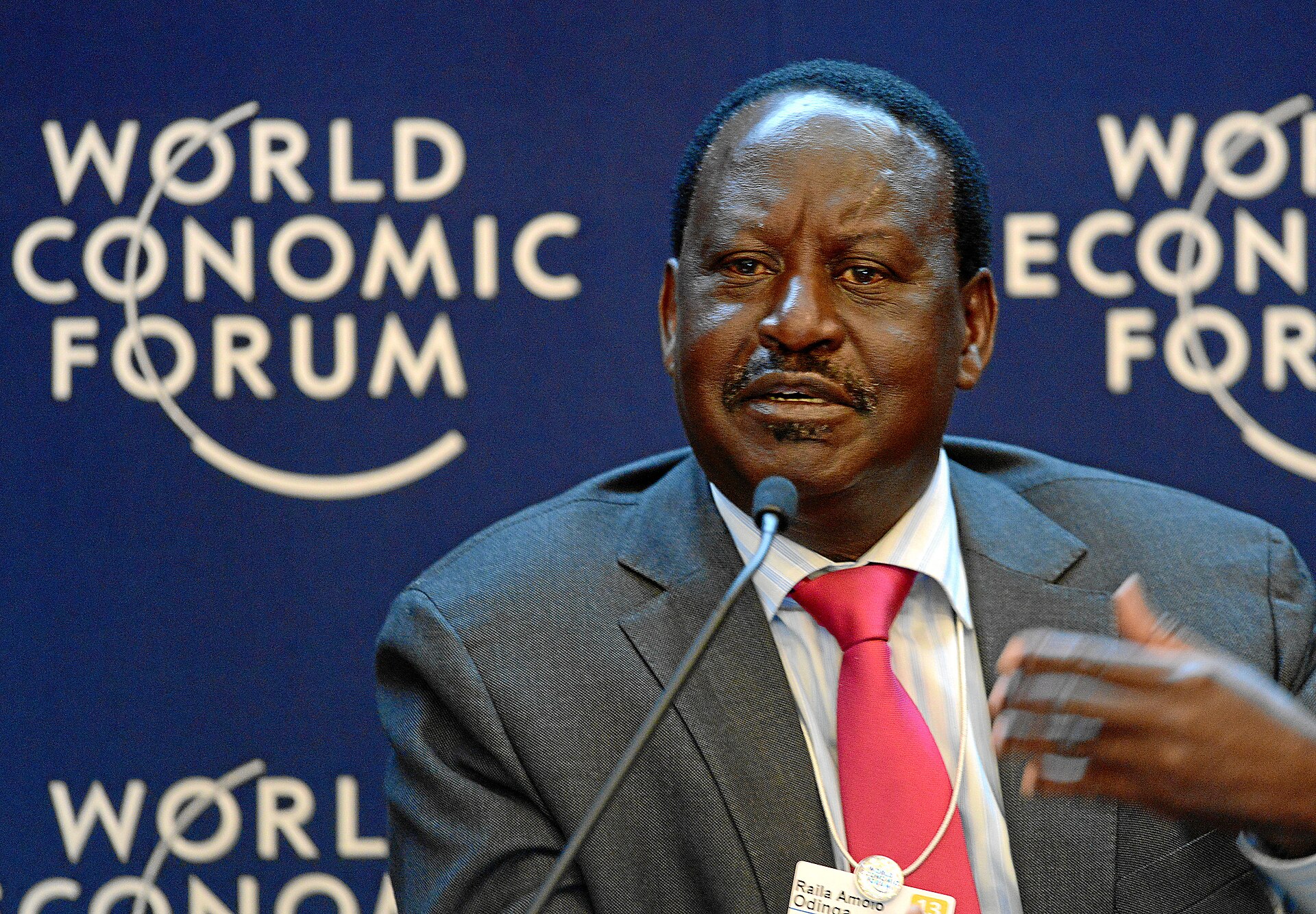 Raila Odinga in a grey suit and red tie speaking into a microphone in front of a blue World Economic Forum backdrop