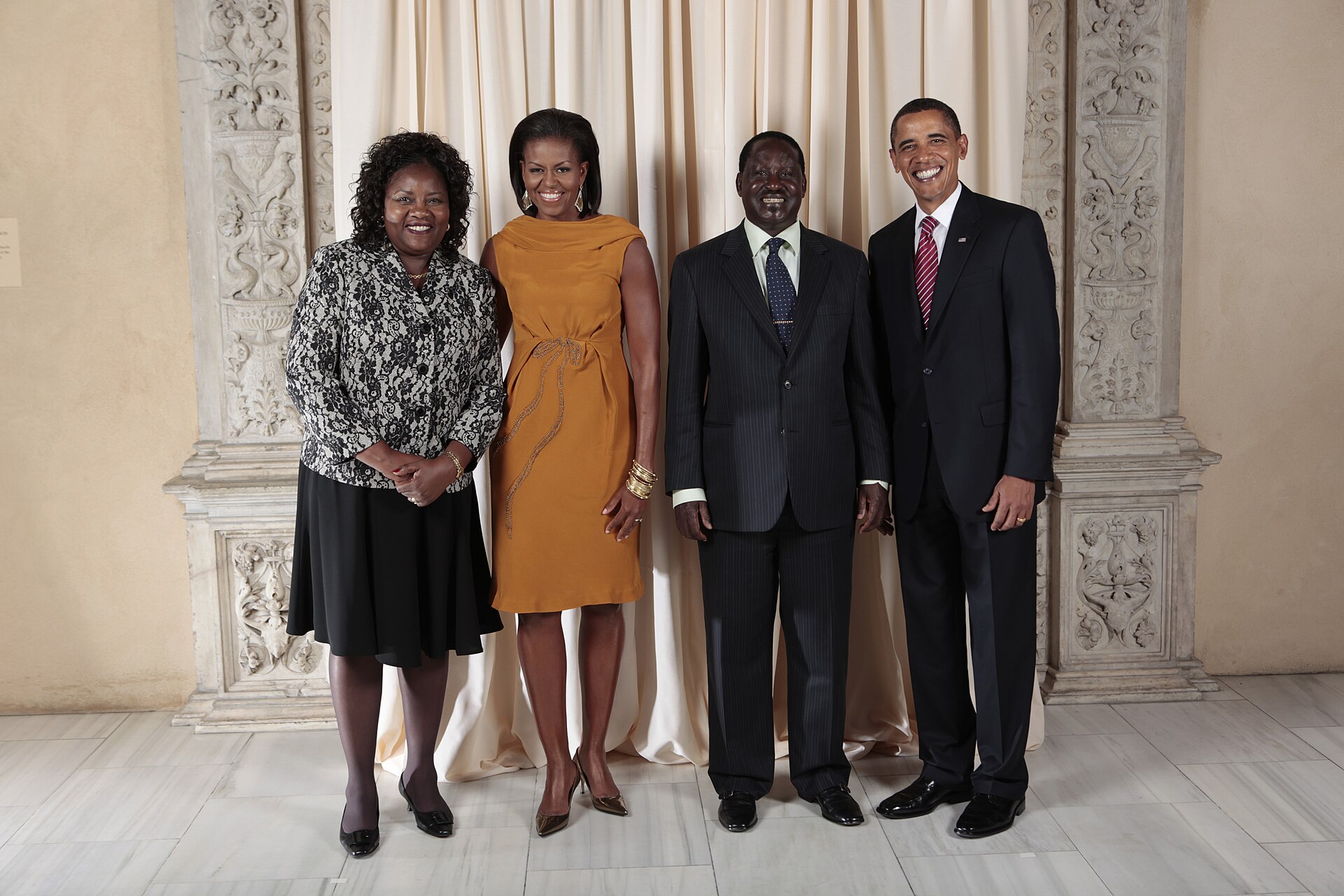 Ida Odinga, Michelle Obama, Raila Odinga and Barack Obama standing side by side in front of a cream curtain and carved stone pillars