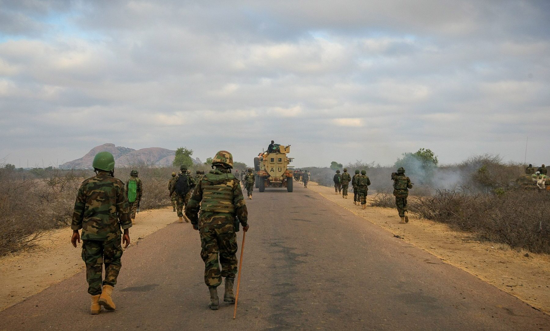 Soldiers in camouflage walk along a paved road through dry scrubland toward a rocky hill, with an armoured vehicle further ahead