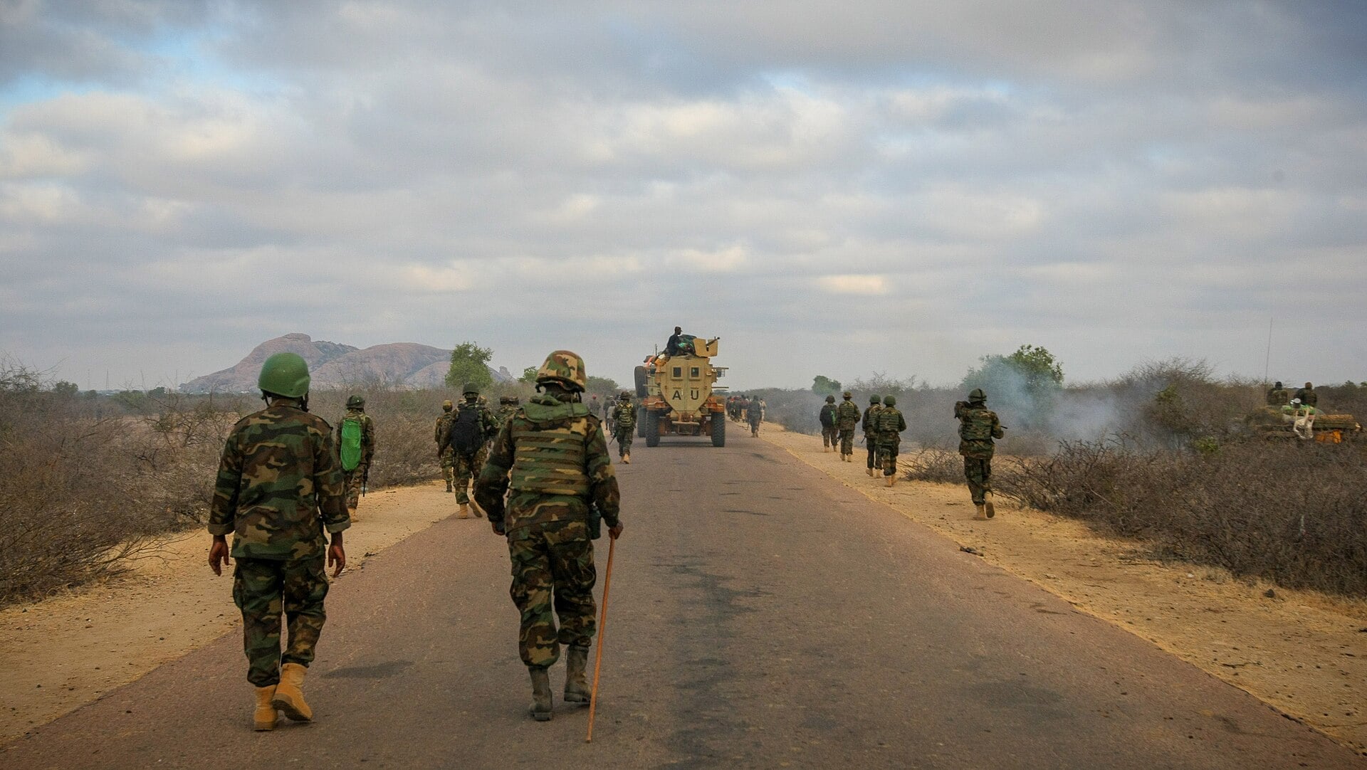 Soldiers in camouflage walk along a paved road through dry scrubland toward a rocky hill, with an armoured vehicle further ahead