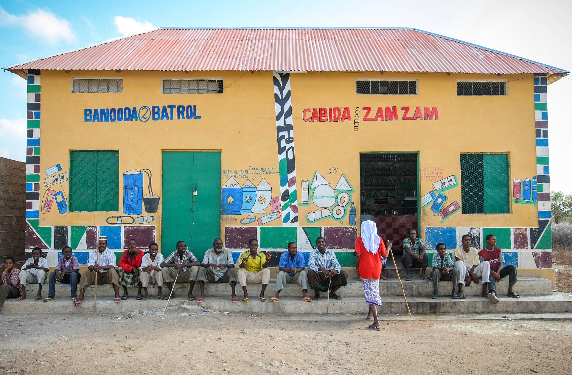 Men sit on the steps of a yellow shop building with painted signs and a red corrugated roof as a woman in a white headscarf walks past