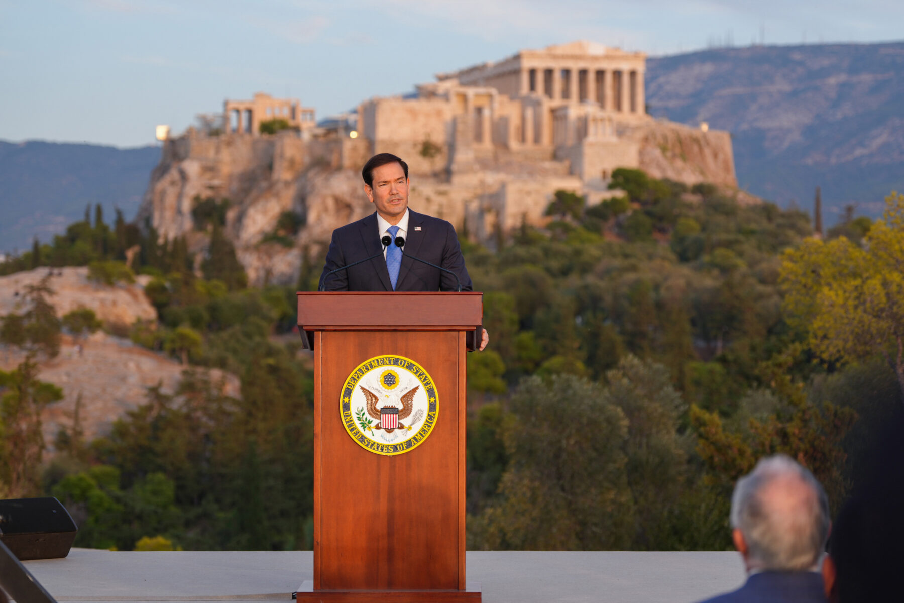 Marco Rubio speaking at a lectern on the Pnyx in Athens with the Acropolis behind him
