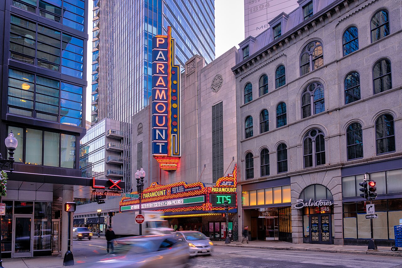 The lit vertical Paramount sign and marquee on Washington Street in Boston at dusk, between glass towers and an old granite building