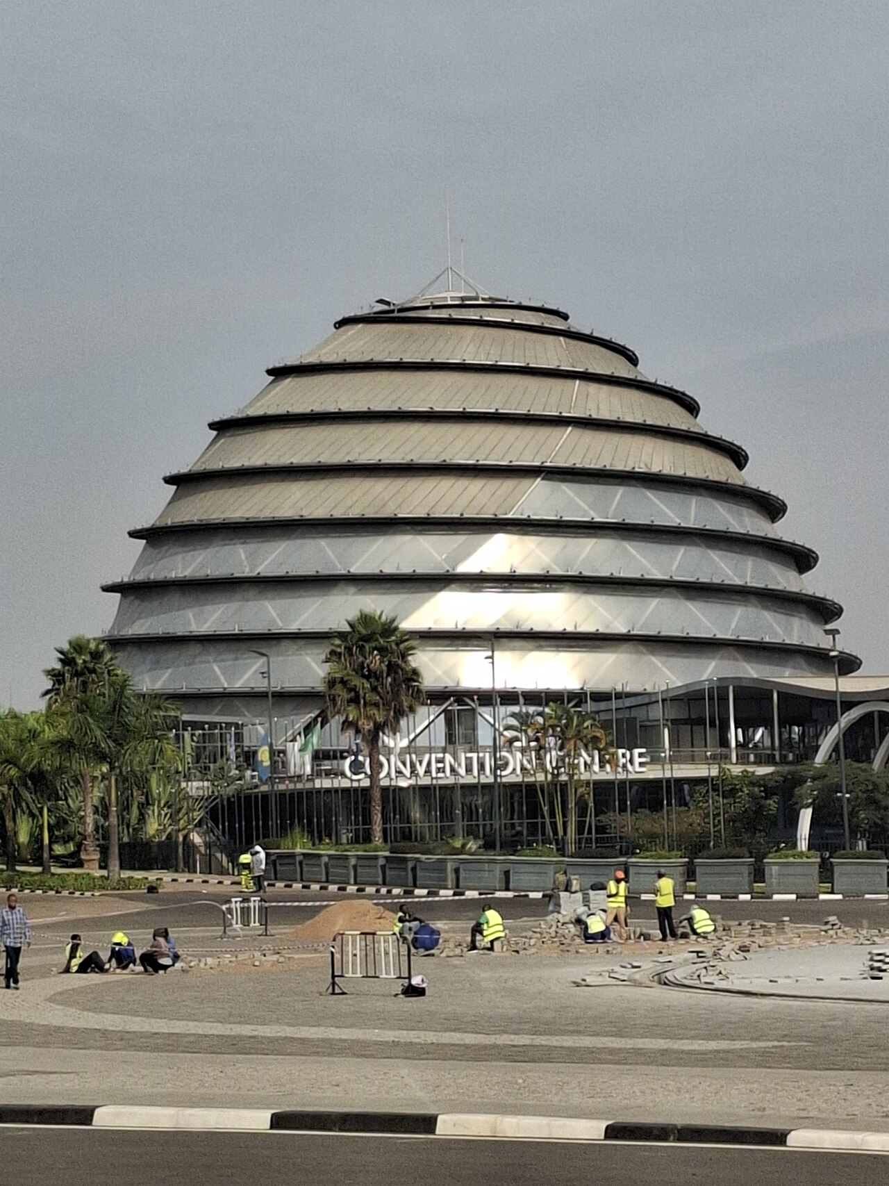 The domed glass-and-metal Kigali Convention Centre, with road workers laying paving stones in the square in front of it.