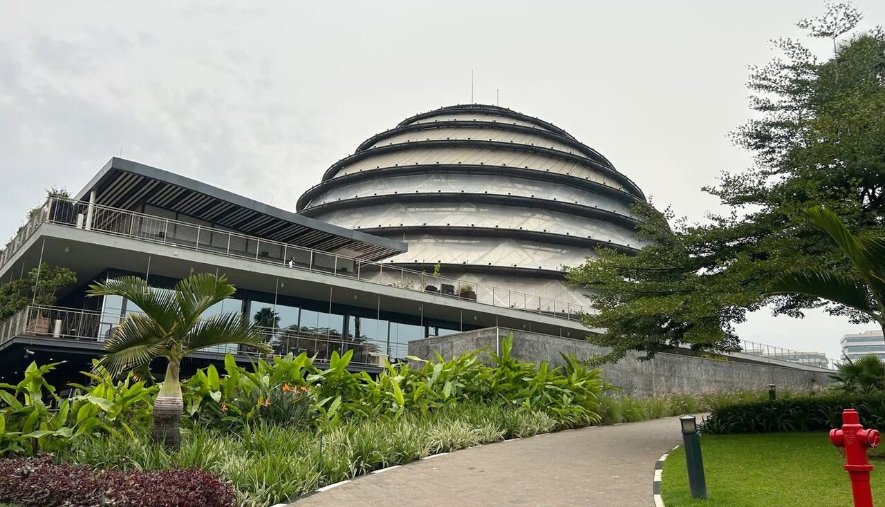 The domed glass-and-steel roof of the Kigali Convention Centre behind a curved terrace, palms and a garden path.