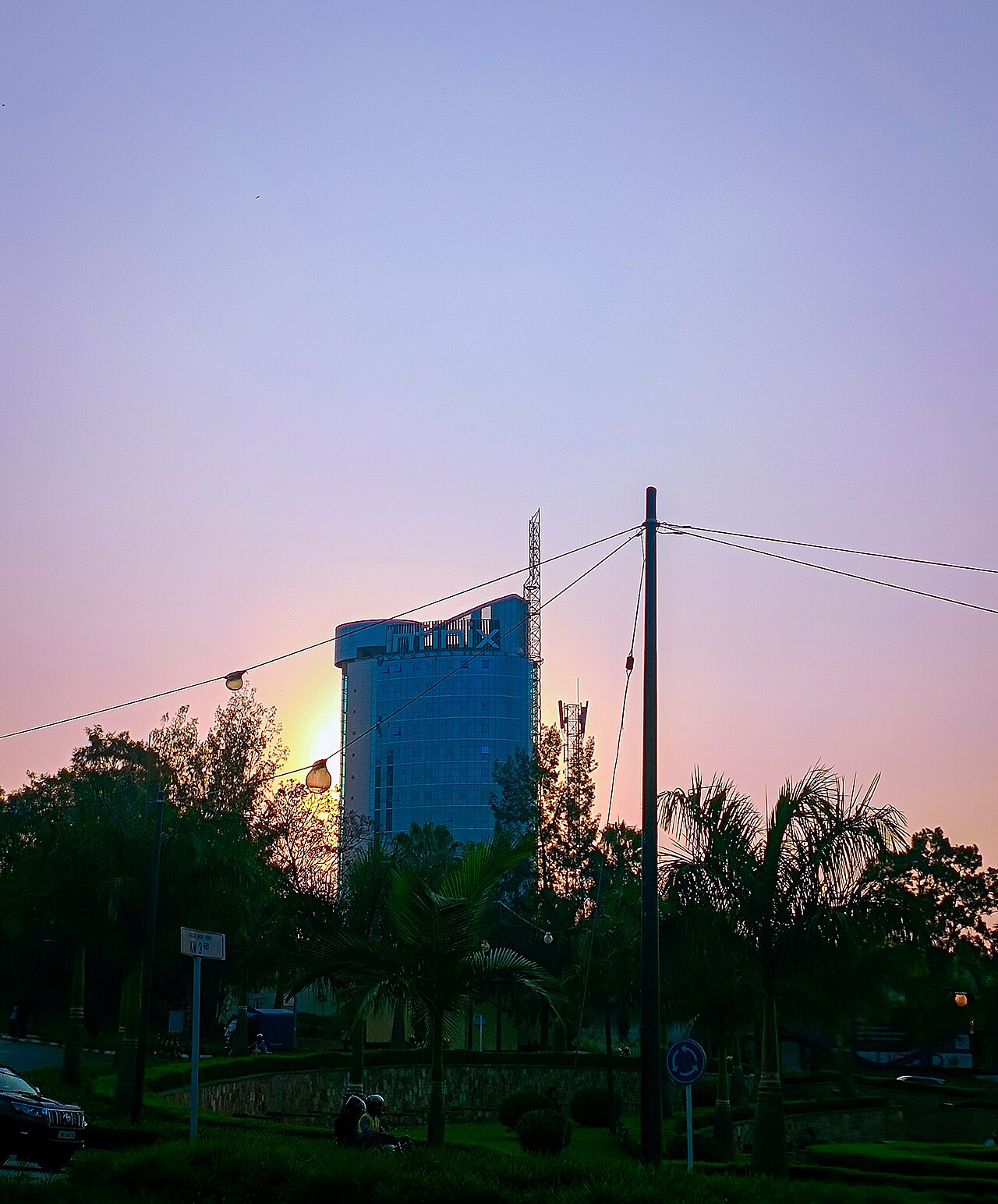 A glass office tower in Kigali against a pink and purple sunset sky, seen between palm trees and street cables.