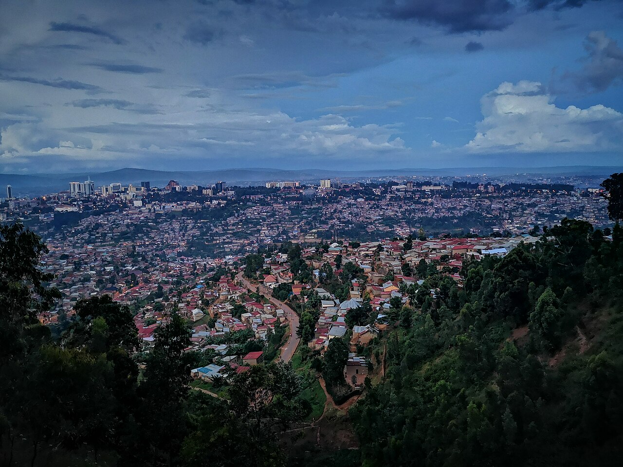 Evening view over the hills of Kigali, with red-roofed homes in the foreground and the towers of the city centre under a cloudy sky.