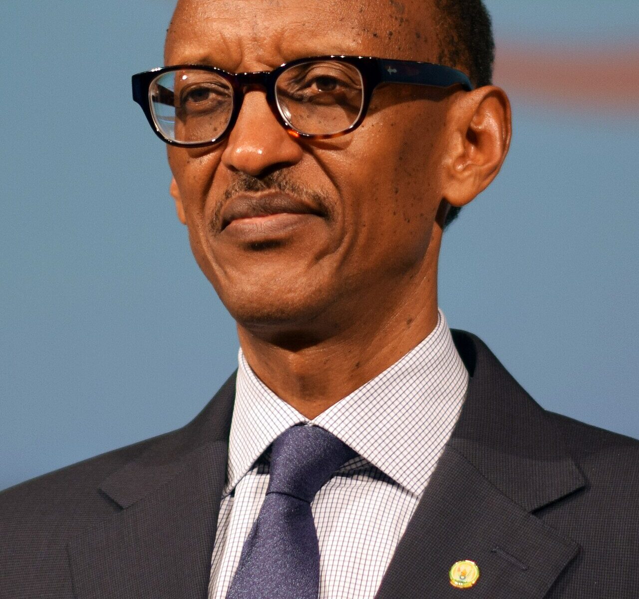 Paul Kagame, President of Rwanda, in a dark suit and blue tie, glasses on, looking to the side
