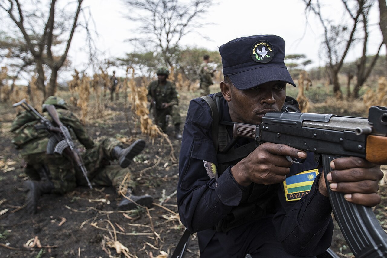 A Rwanda National Police officer aims a rifle during a field exercise, with Rwandan soldiers behind him