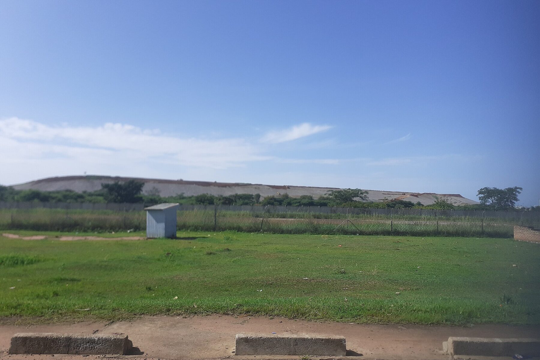 Grassy roadside field with a small metal hut in front of the long, flat waste heap of the Buzwagi gold mine under a blue sky