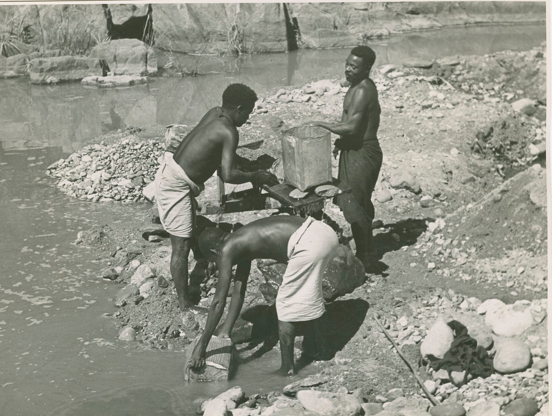 Black-and-white photo of three men washing and sifting gravel with a metal box and tools at the edge of a shallow river
