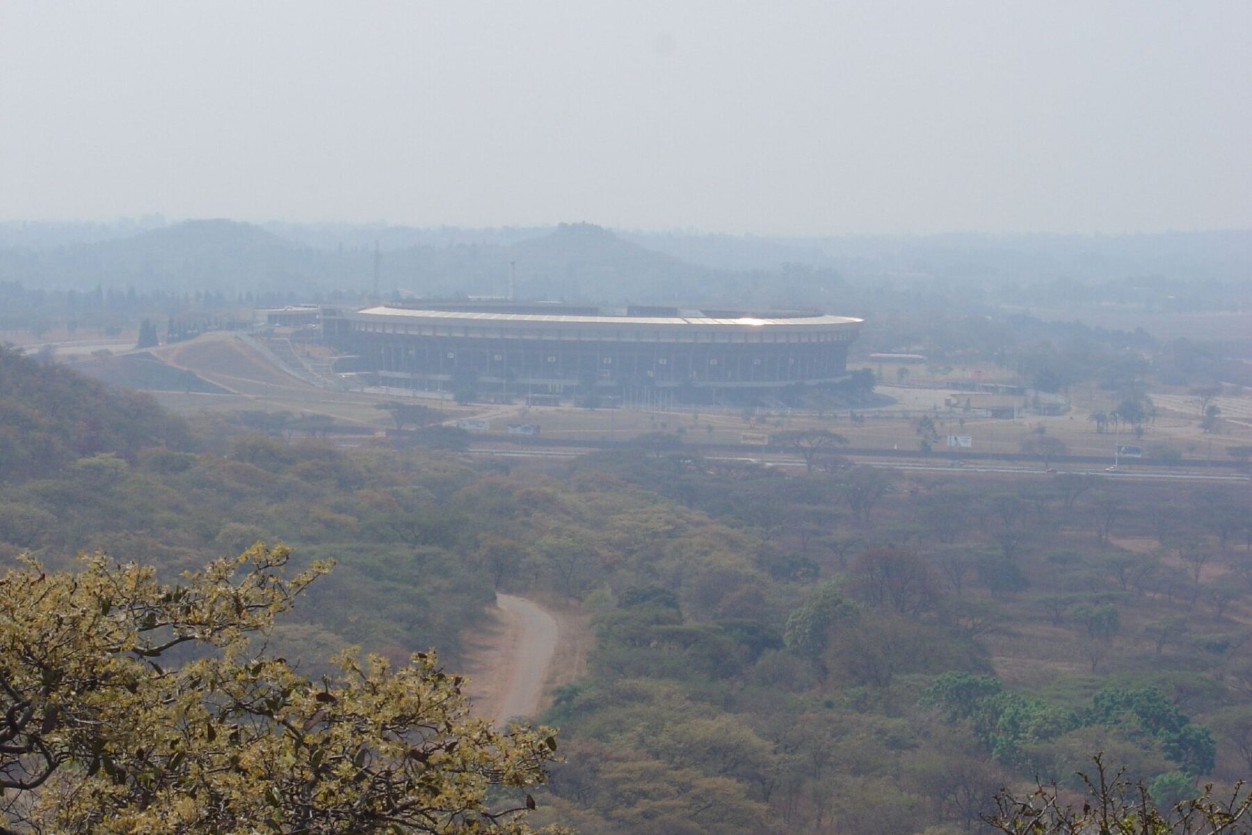 A large oval stadium on open ground below wooded hills, seen from a hillside in hazy light