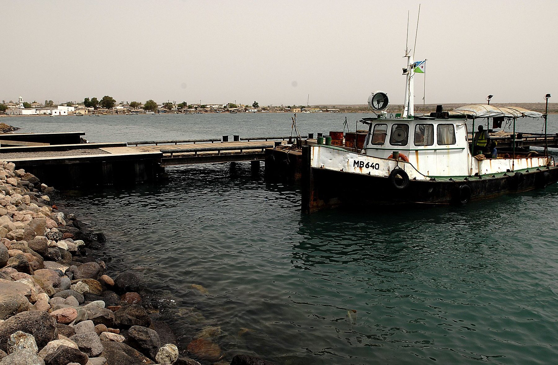 A white and black tugboat flying the Djibouti flag moored at a pier in Obock harbour, with rocks in the foreground and the low town on the far shore