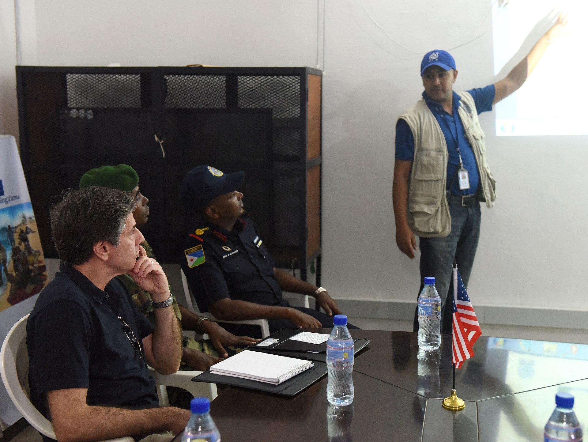 A man in an IOM cap and vest points at a projected screen while a US official and two Djiboutian officers in uniform listen at a table with a small US flag