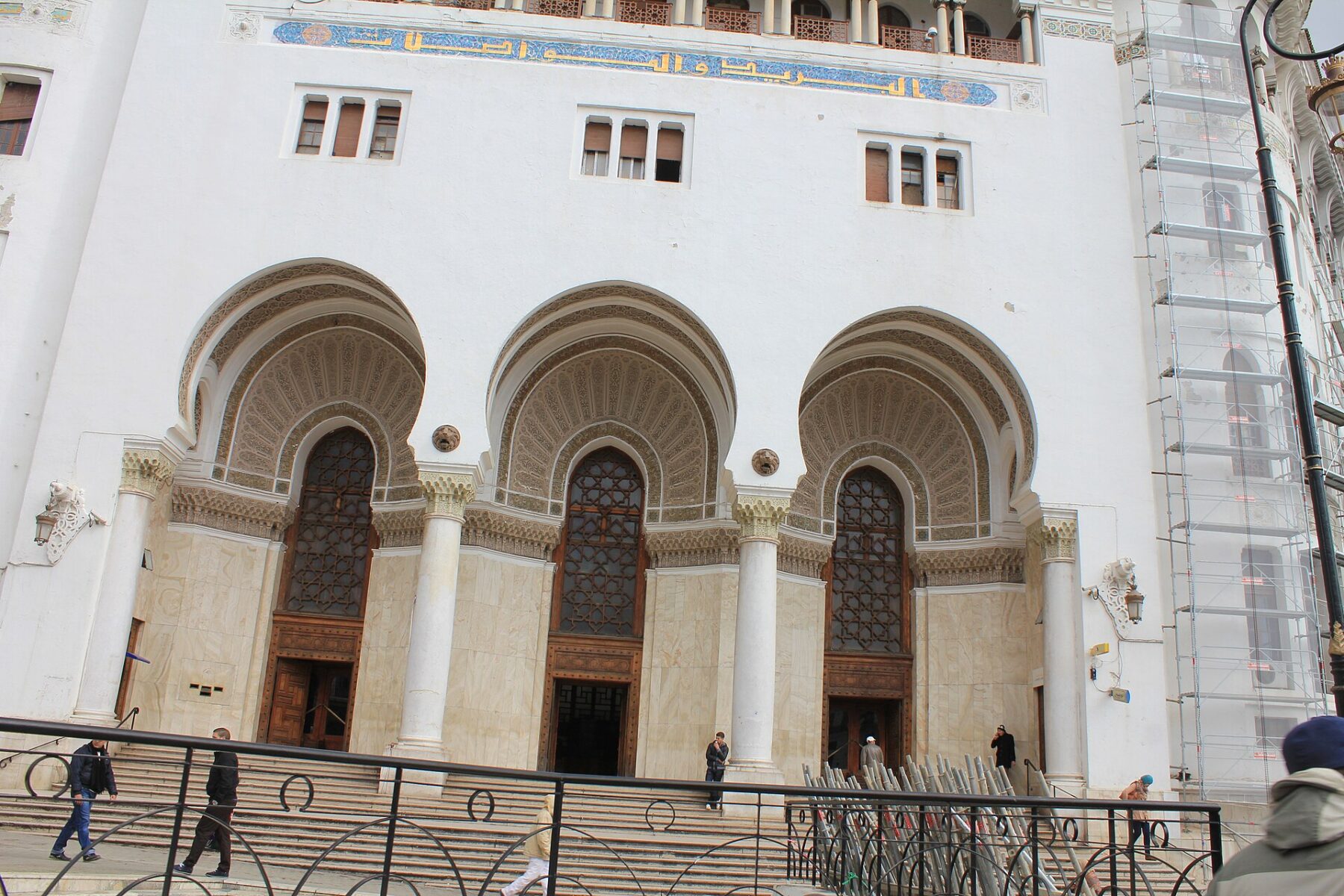 The white arched facade and main entrance stairs of the Grande Poste building in central Algiers, with pedestrians and scaffolding on one side