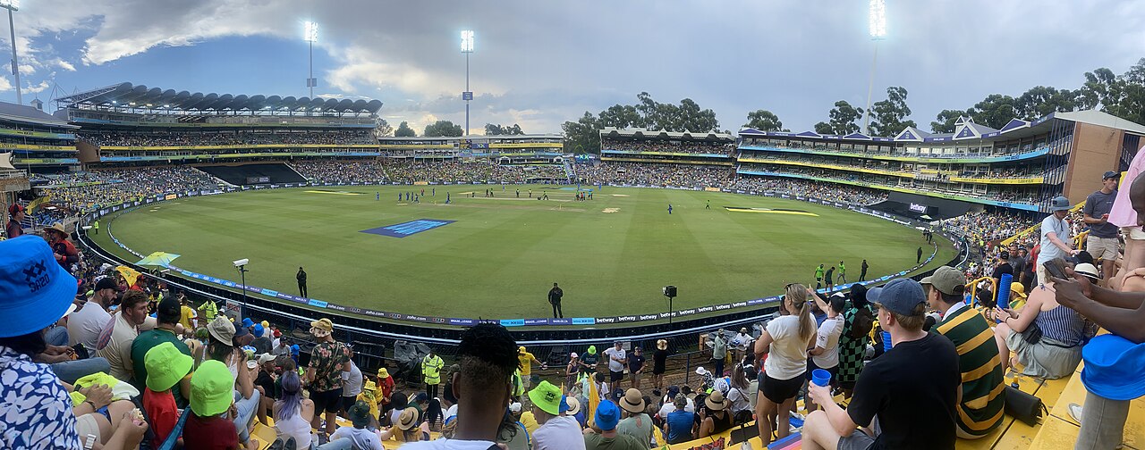 Panoramic view of the Wanderers cricket stadium in Johannesburg during an SA20 match, with spectators in sun hats in the foreground, floodlights and packed stands around the green field.