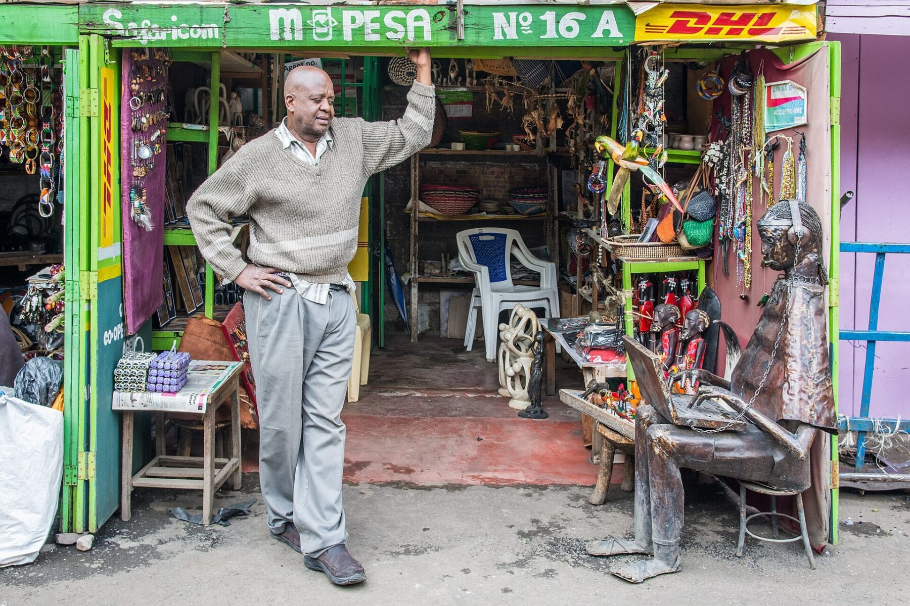 A man in a grey sweater leans in the doorway of a green Safaricom M-Pesa agent shop selling crafts in Nairobi