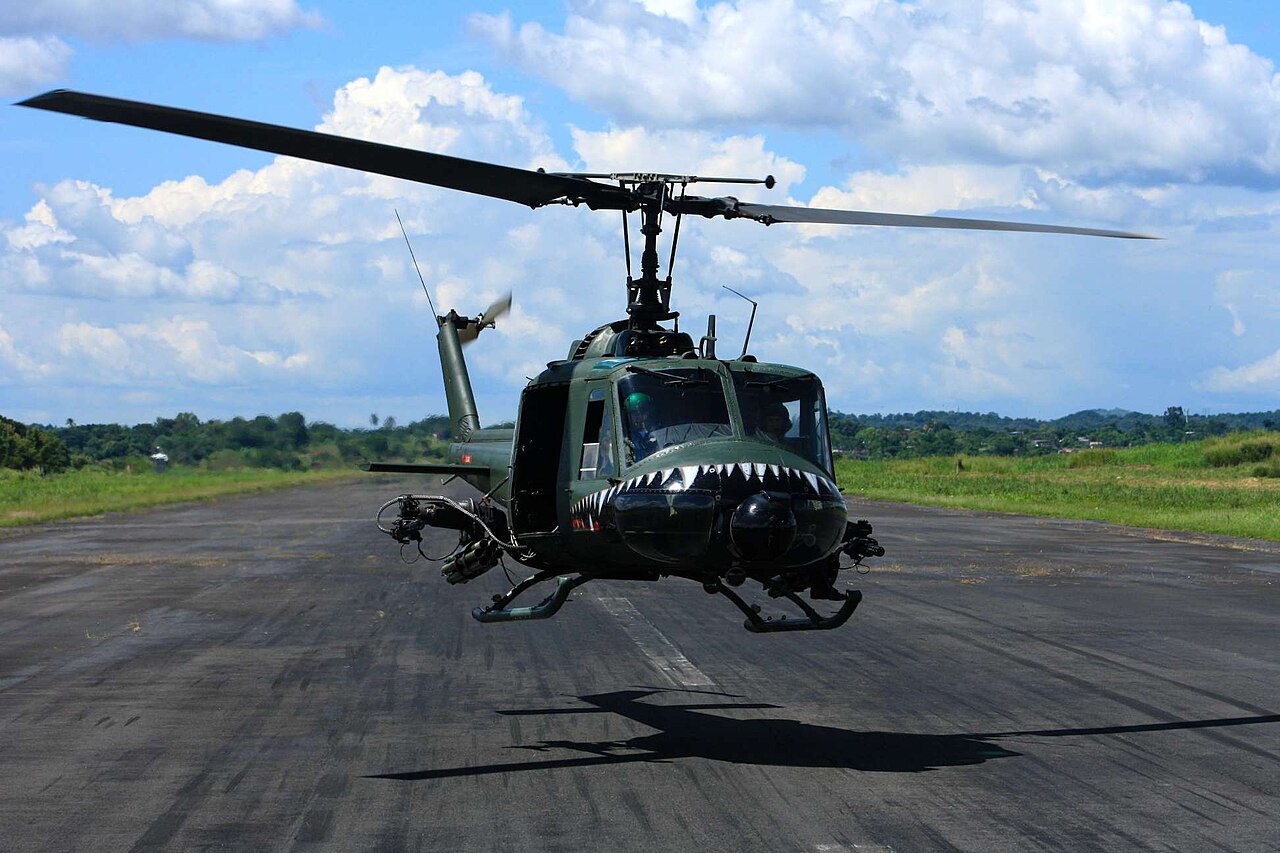 A green Bell UH-1 helicopter with a shark-mouth nose hovering above a runway
