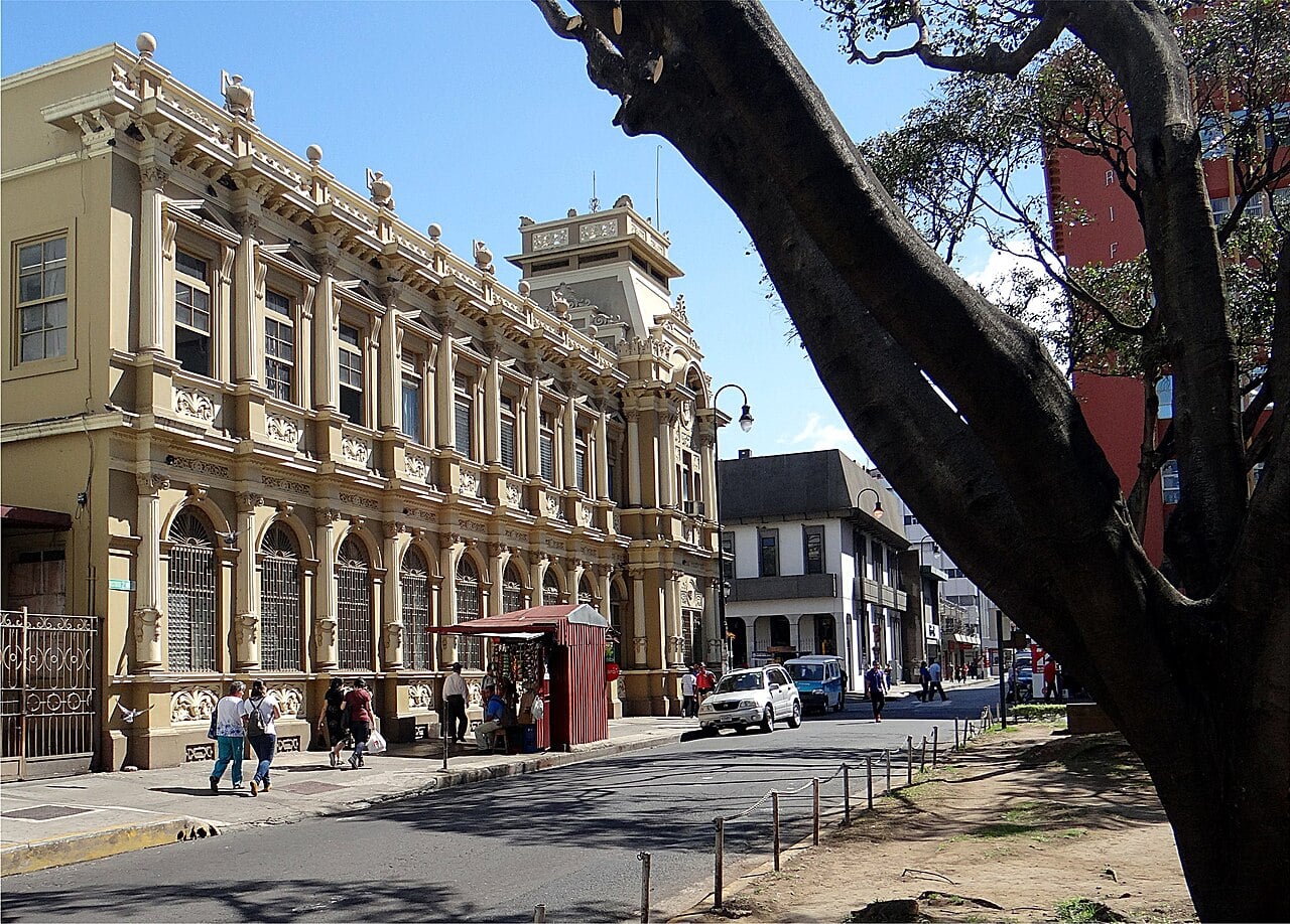 A cream-colored historic building with arched windows on a sunny street in central San José, Costa Rica, with pedestrians and a street kiosk.