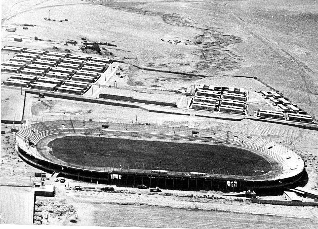 Black-and-white aerial photograph of the Estadio Carlos Dittborn in Arica, Chile, a large oval stadium surrounded by desert and housing blocks