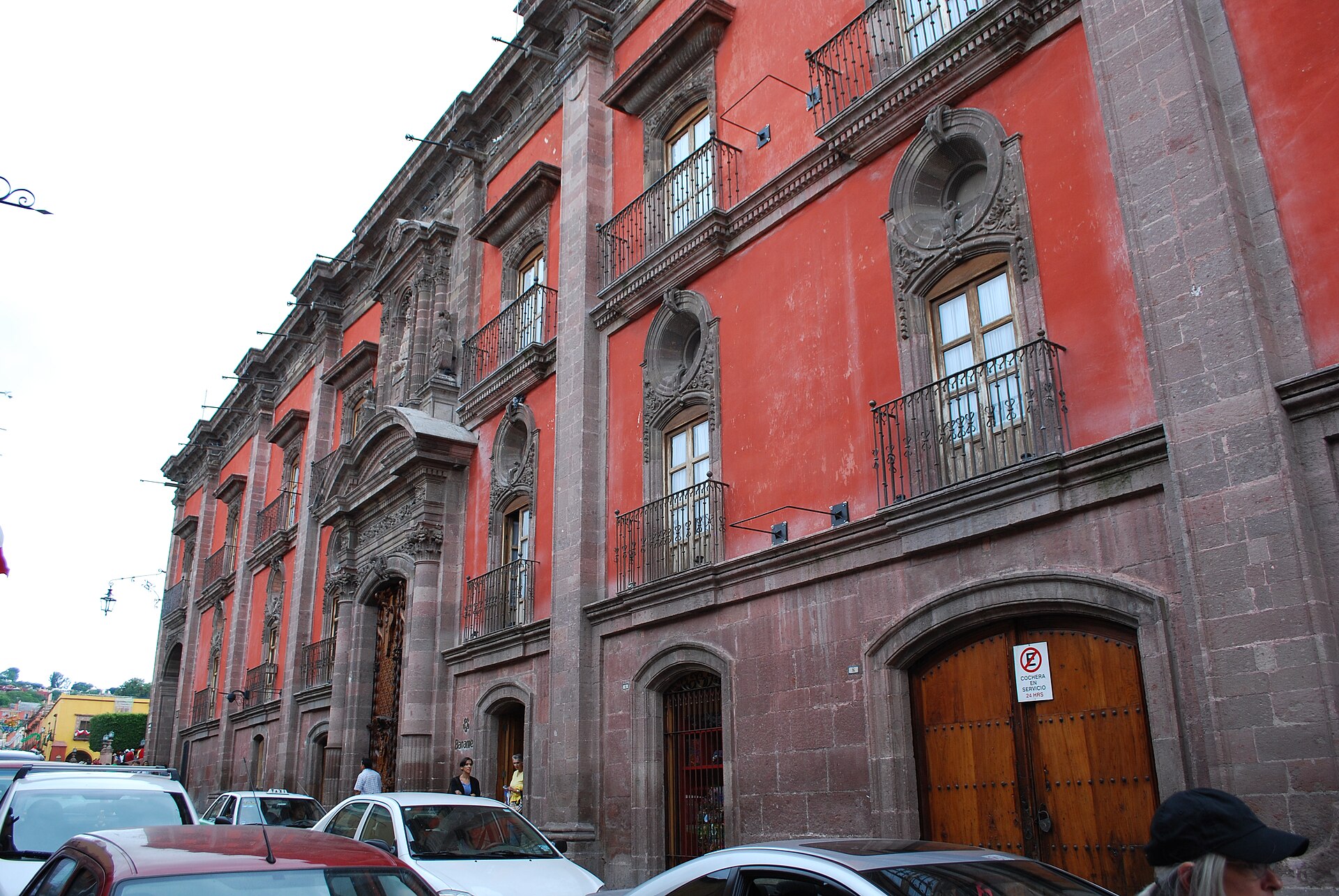 Red colonial mansion with carved stone window frames, iron balconies and a large wooden door, with cars parked along the street in San Miguel de Allende