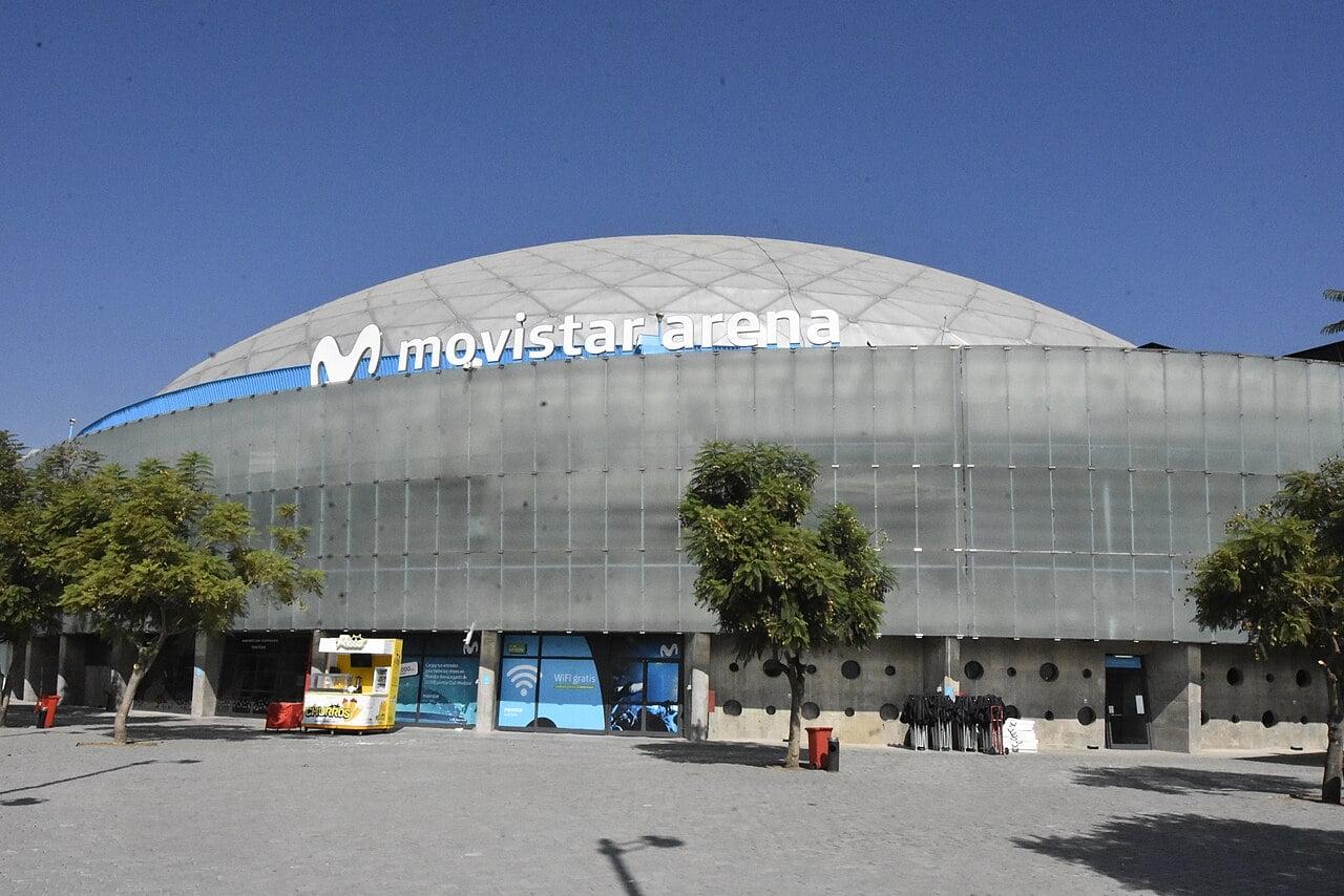 Exterior of the domed Movistar Arena in Parque O'Higgins, Santiago, under a clear blue sky, with trees and a snack kiosk at the entrance.