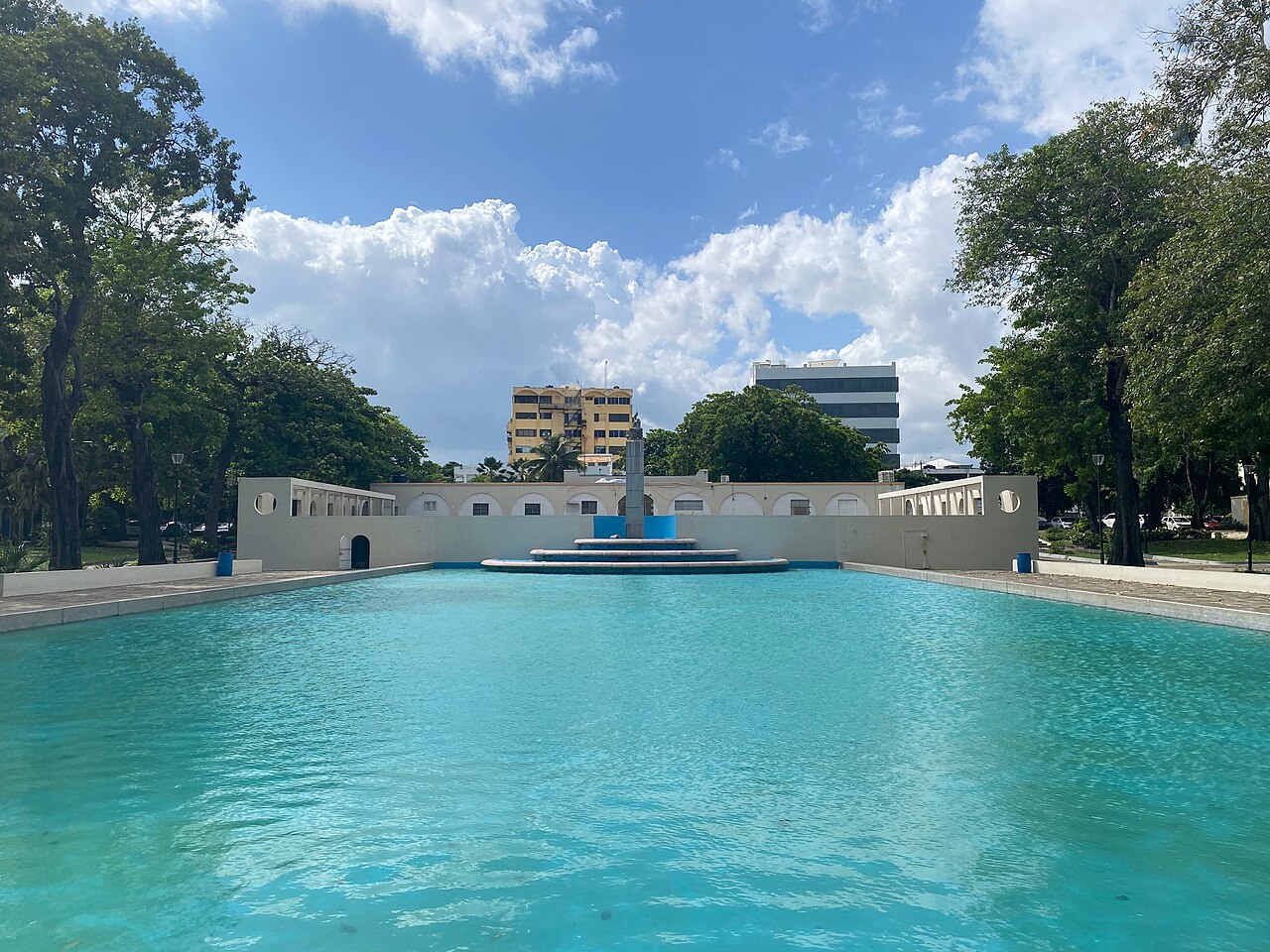 Turquoise reflecting pool with a central monument in Parque Eugenio María de Hostos, Santo Domingo, under a blue sky with clouds.