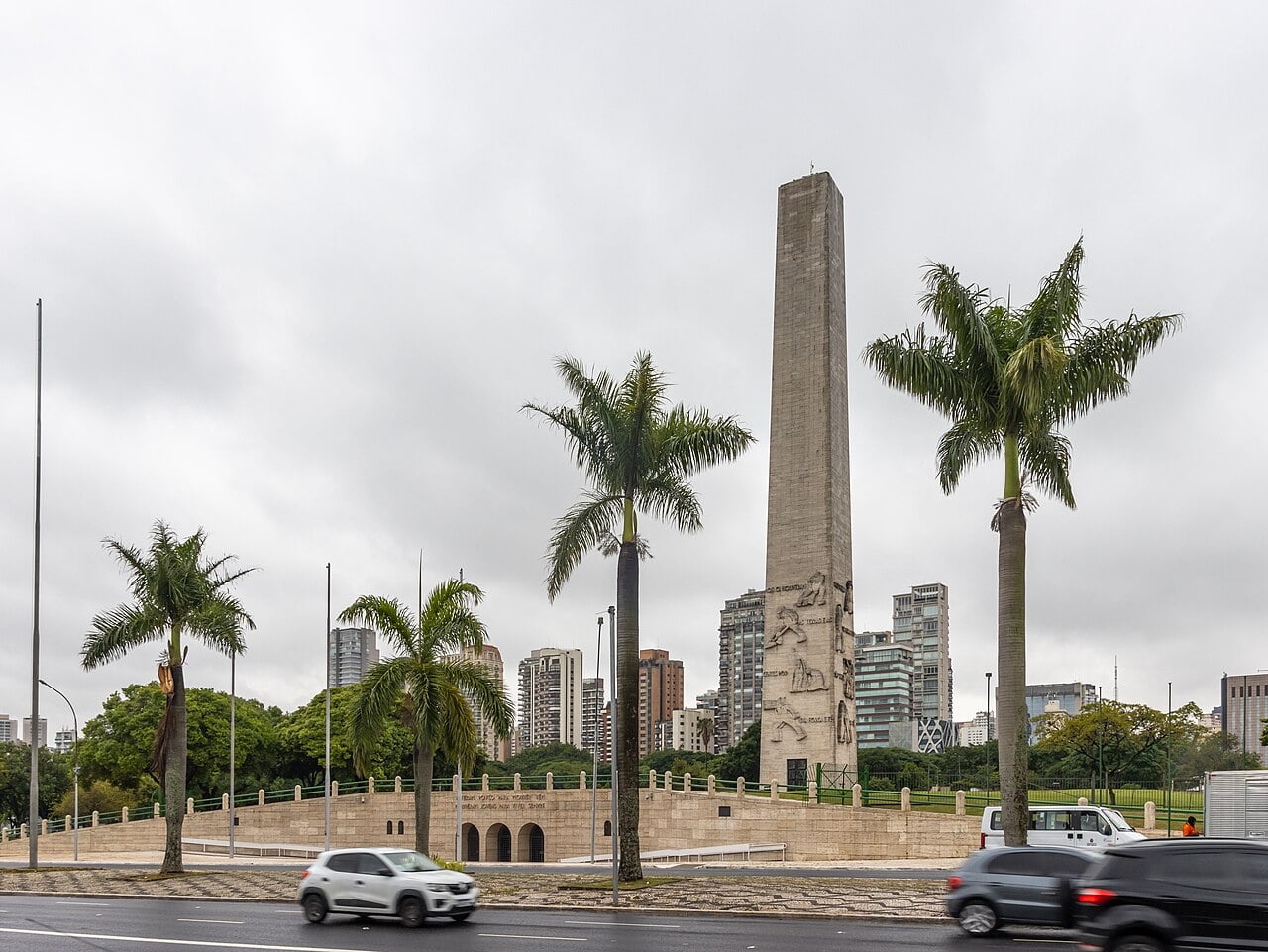 A tall stone obelisk with carved reliefs stands behind royal palm trees at Ibirapuera in São Paulo, with apartment towers behind it under a grey sky.