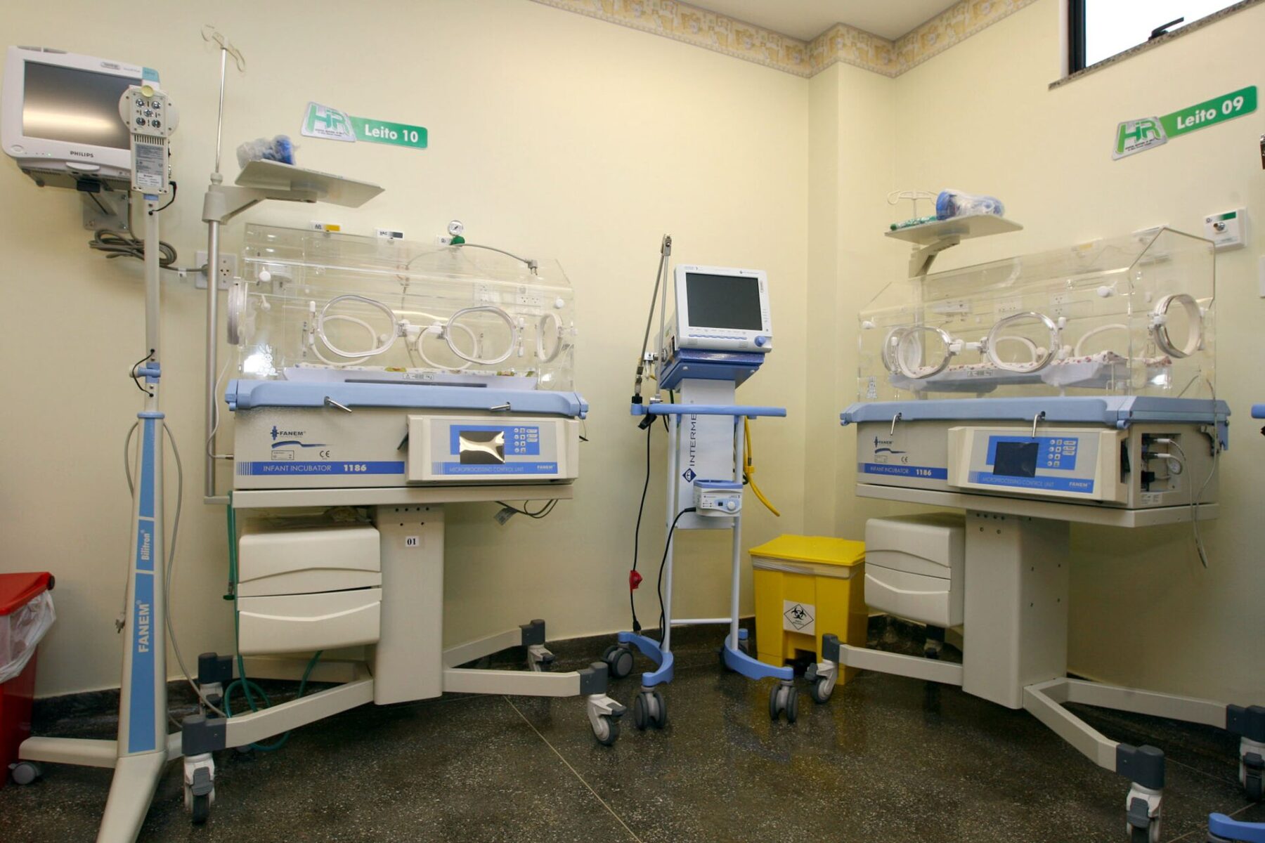 Two empty infant incubators with monitors and bed number labels in a neonatal intensive care room with yellow walls
