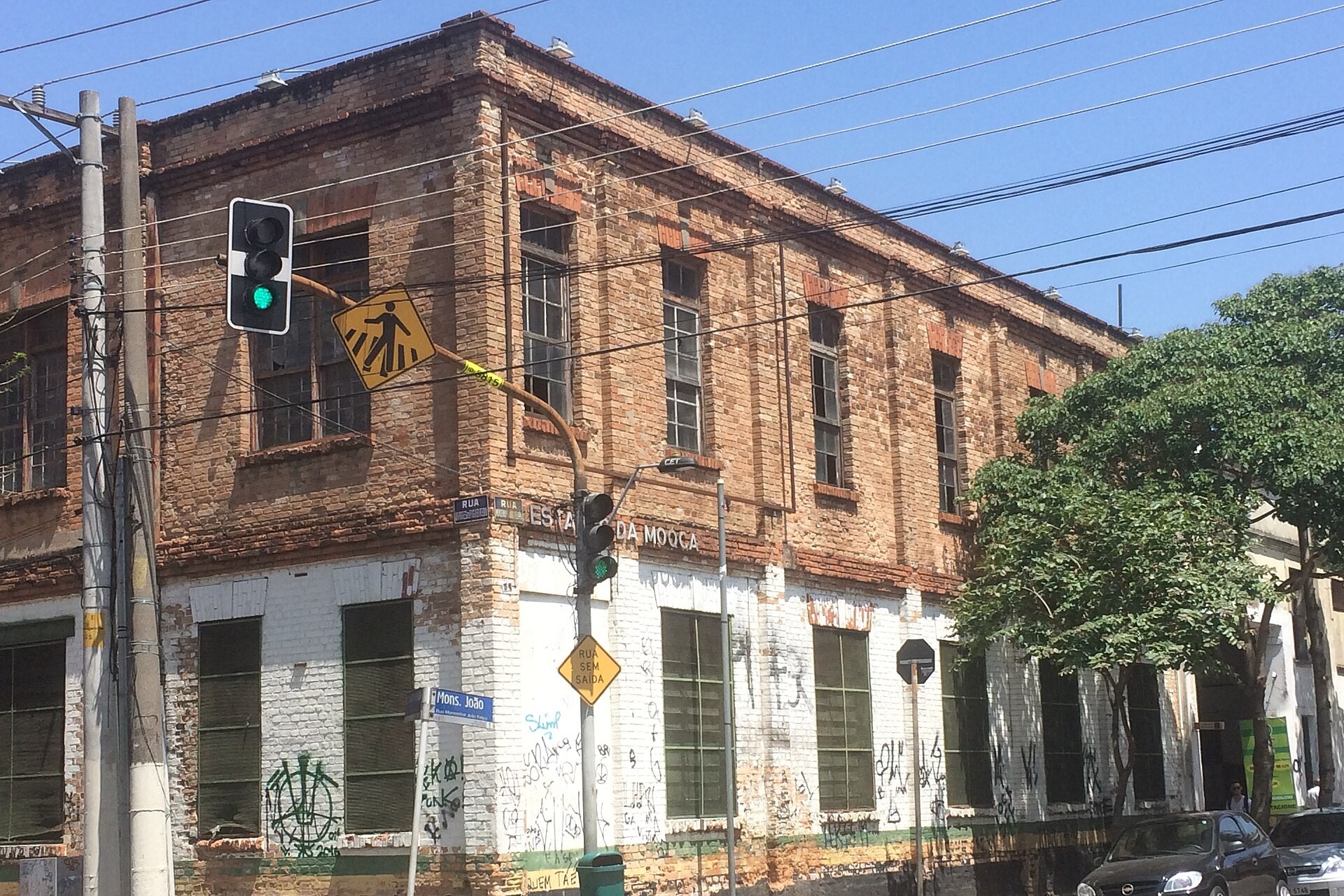 Corner of a two-storey brick building with a pedestrian-crossing sign and traffic lights under a blue sky on a street in the Mooca district of São Paulo