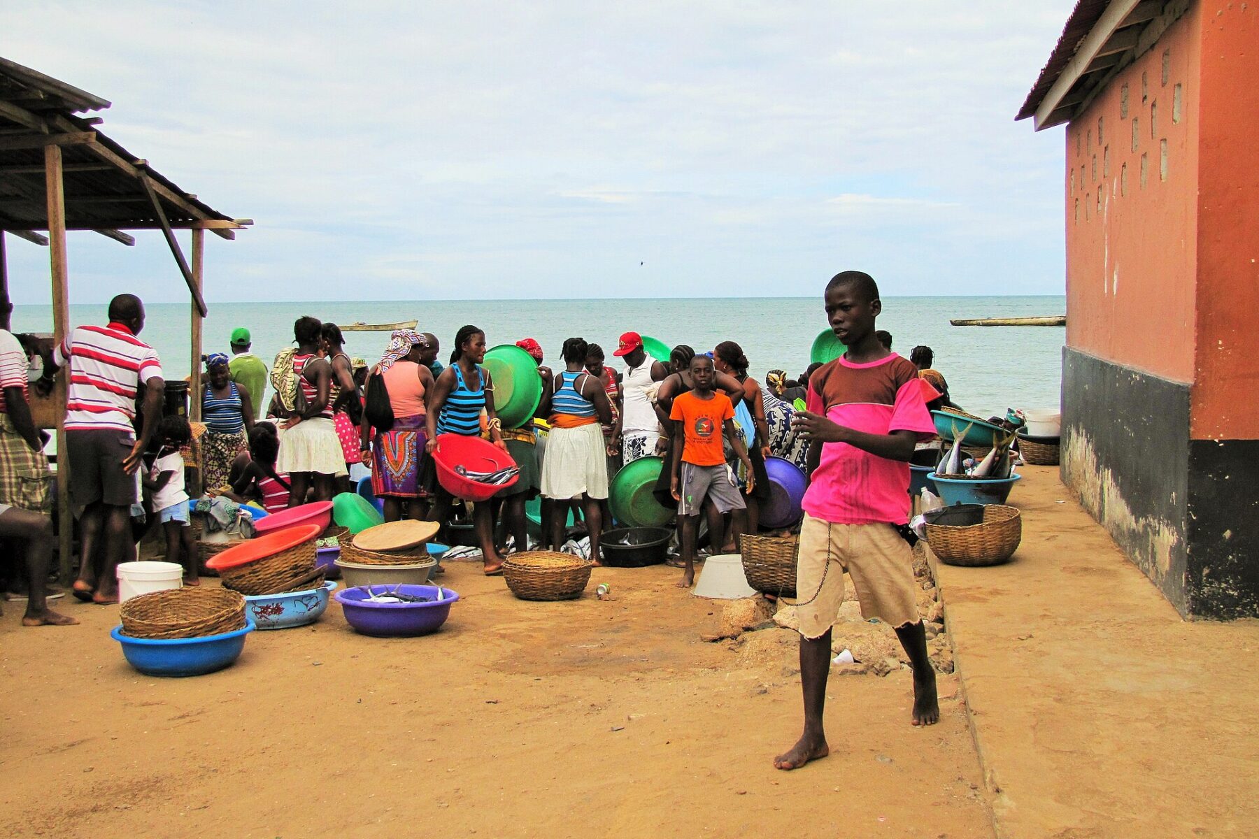 Women and children sell fish from colourful basins on a beach beside a pink building in São Tomé