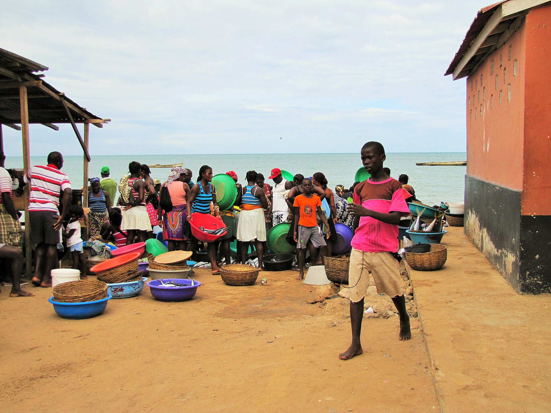 Women and children sell fish from colourful basins on a sandy beach beside an orange building in São Tomé