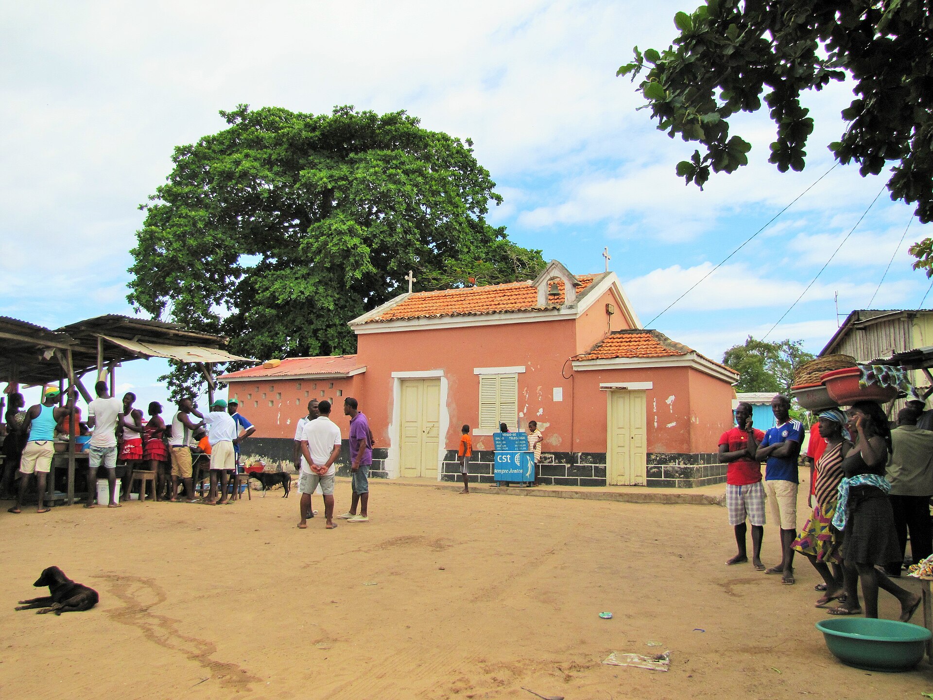 Villagers stand in a sandy square in front of a small pink chapel with a tiled roof and a large tree in São Tomé