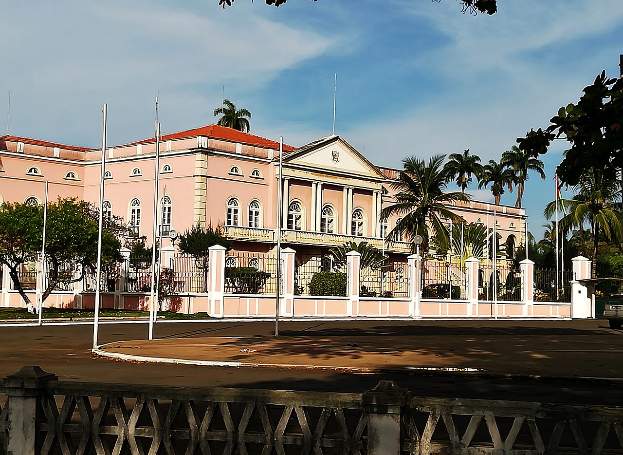 Pink presidential palace in São Tomé seen from the street, with a red roof, white gate posts and palm trees